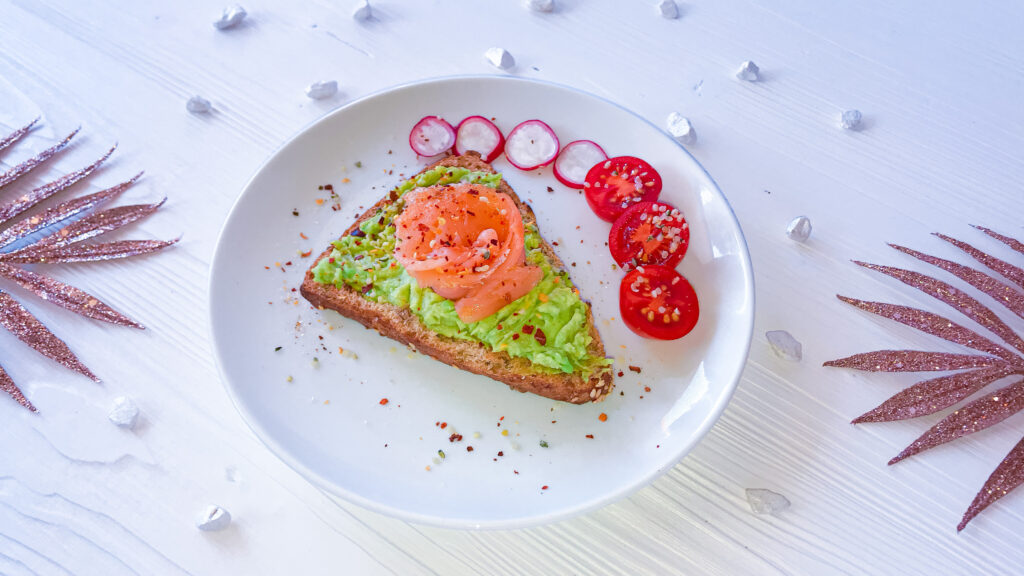 Gesunder Avocado-Toast mit Lachs, Radischen- und Tomatenscheiben mit Chilisalz bestreut und auf einem weißen Teller angerichtet. 