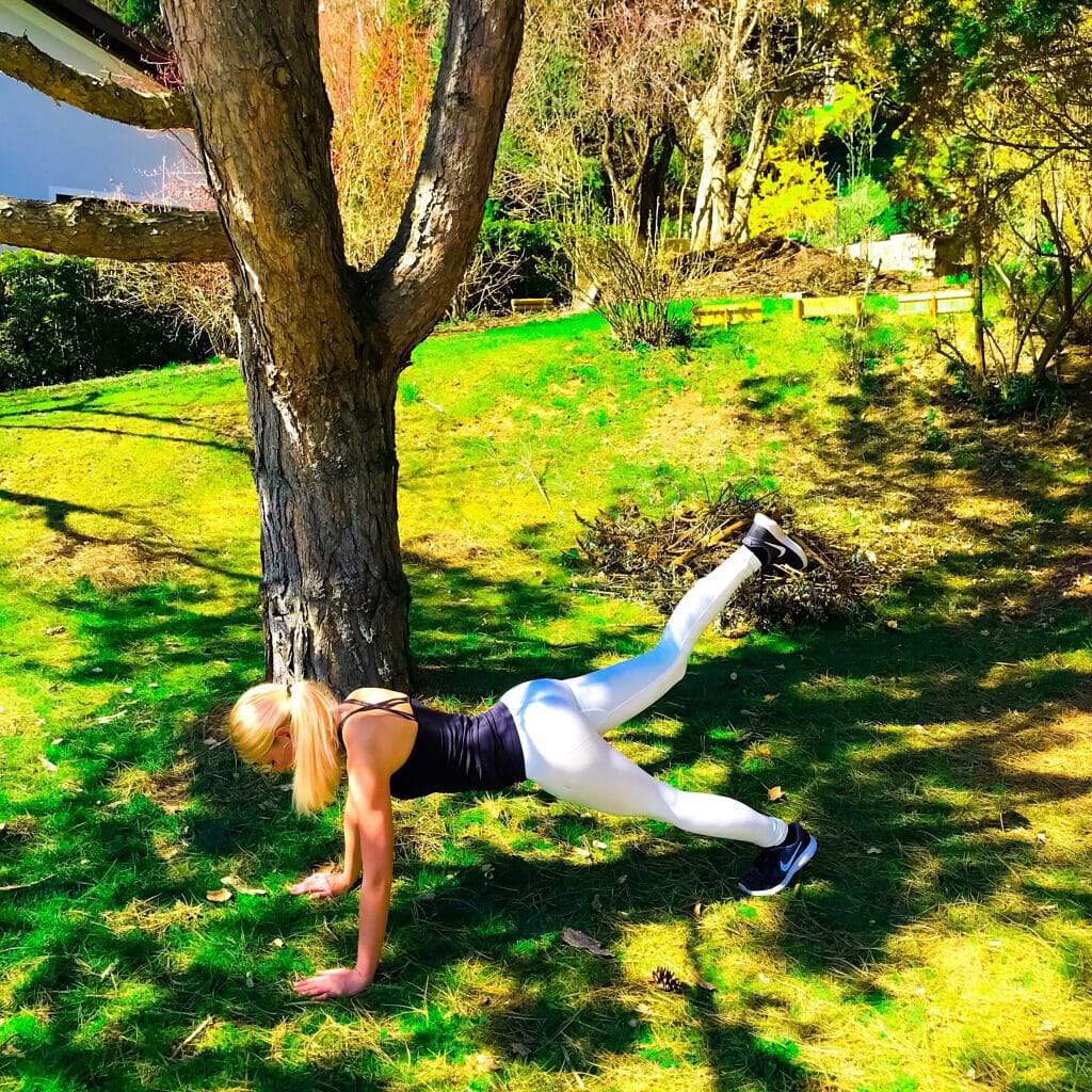Fitnessmädchen im Garten in Stützposition mit einem scharzen Top und weißen Fitnessleggings. 