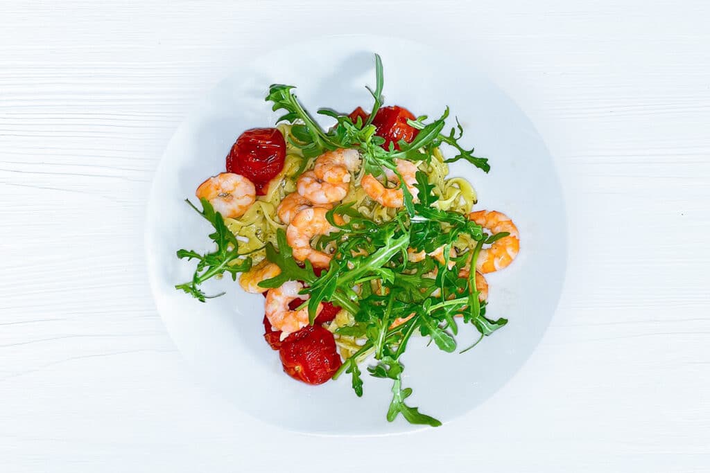 Tagliatelle mit Rucola-Pesto, Ofentomaten und Garnelen in einer weißen Schale serviert