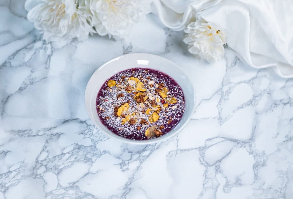 Gesunde vegane Smoothie Bowl aus Beeren garniert mit Granola und Kokosflocken in einer weißen Schale auf einem Marmortisch serviert. 