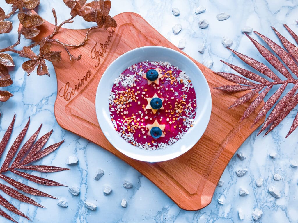 Beeren-Smoothie-Bowl in einer weißen Schale serviert mit Bluetenpollen, Kokosflocken, Heidelbeeren und Bananenscheiben. 