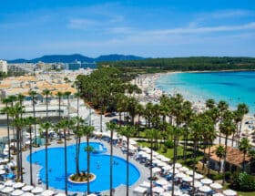 Aussicht vom Dach auf den großen hoteleigenen Pool auf Mallorca Sa Coma umgeben von hohen Palmen. Im Hintergrund sieht man das türkisfarbene Meer und den weißen Sandstrand. bei schöne Wetter.
